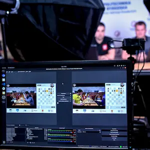 Monitor komputerowy z uruchomionym programem OBS Studio do transmisji szachów na żywo, wyświetlający widok na dwie partie oraz szachownicę cyfrową. W tle widoczne profesjonalne studio nagraniowe z oświetleniem i komentatorami.