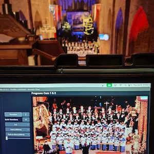 Laptop wyświetlający podgląd transmisji na żywo z występu chóru w katedrze. Na ekranie widać chór w strojach liturgicznych, a w tle rozmazany widok na nawę kościoła z choinkami i ołtarzem.