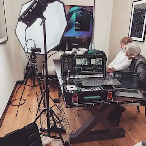 Stanowisko do realizacji transmisji wideo w nowoczesnym pokoju. Przy stole siedzą dwie osoby obsługujące laptopy oraz mikser wideo zamontowany w profesjonalnej walizce typu flight case. Po lewej stronie stoi duża lampa studyjna z softboxem skierowana w stronę planu.