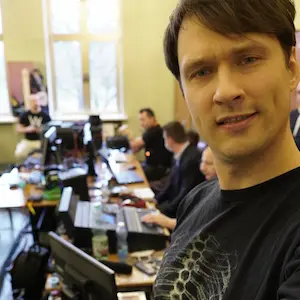 Zdjęcie typu selfie wykonane przez mężczyznę w czarnej koszulce, przedstawiające kulisy realizacji wydarzenia. W tle widać długi stół z licznymi monitorami, mikserami dźwięku i laptopami, przy których pracują inne osoby w słabo oświetlonym pomieszczeniu z dużymi oknami.