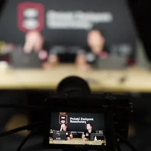 Widok zza kamery Sony na dwóch komentatorów siedzących przy stole na tle ścianki z logo Polskiego Związku Szachowego. Na małym ekranie podglądu aparatu widać ostry obraz mężczyzn, podczas gdy rzeczywista scena w tle jest mocno rozmyta.